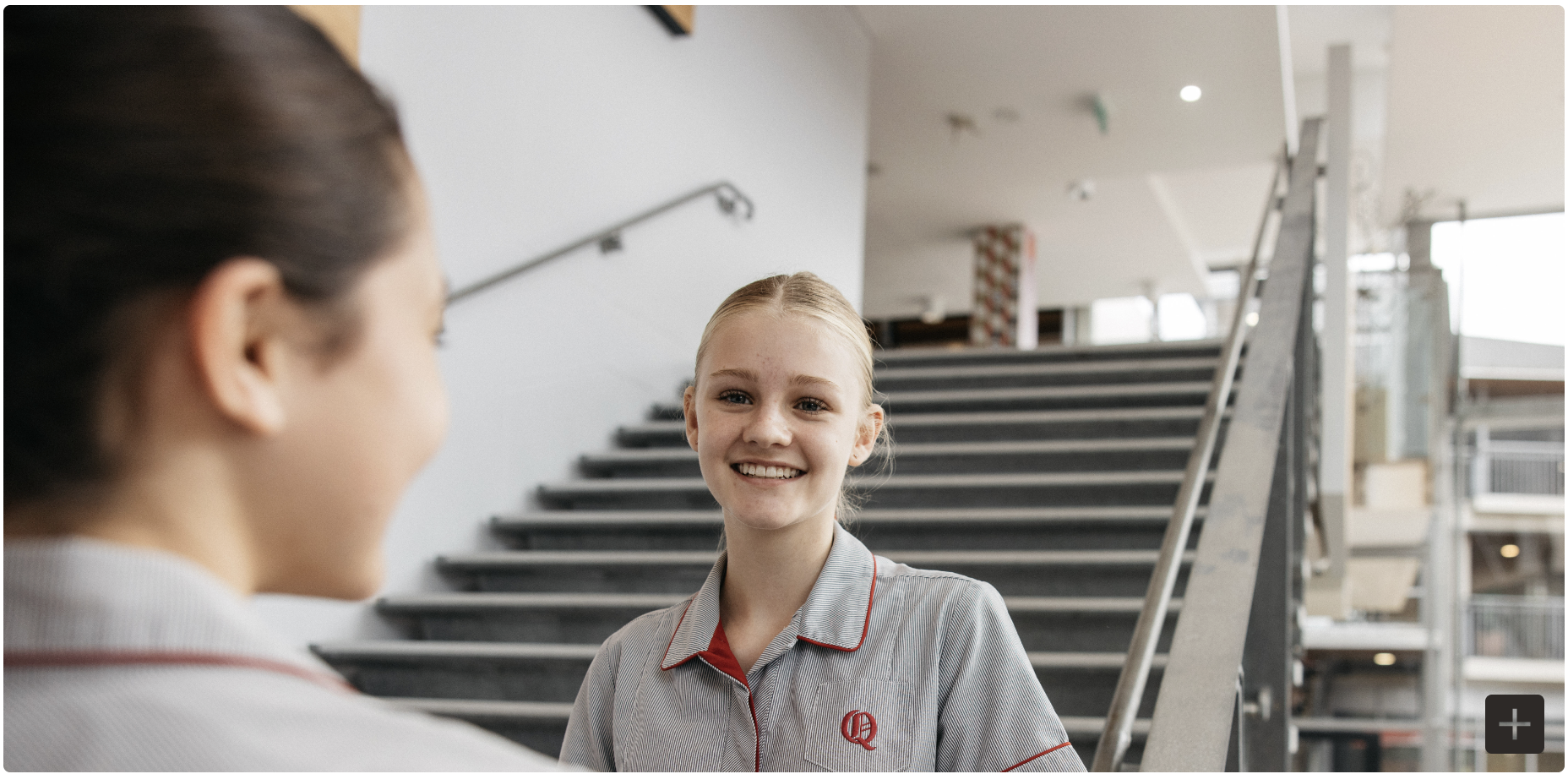 Two Queenwood students in gray and white striped school uniforms with red piping having a conversation on an interior staircase with modern architecture